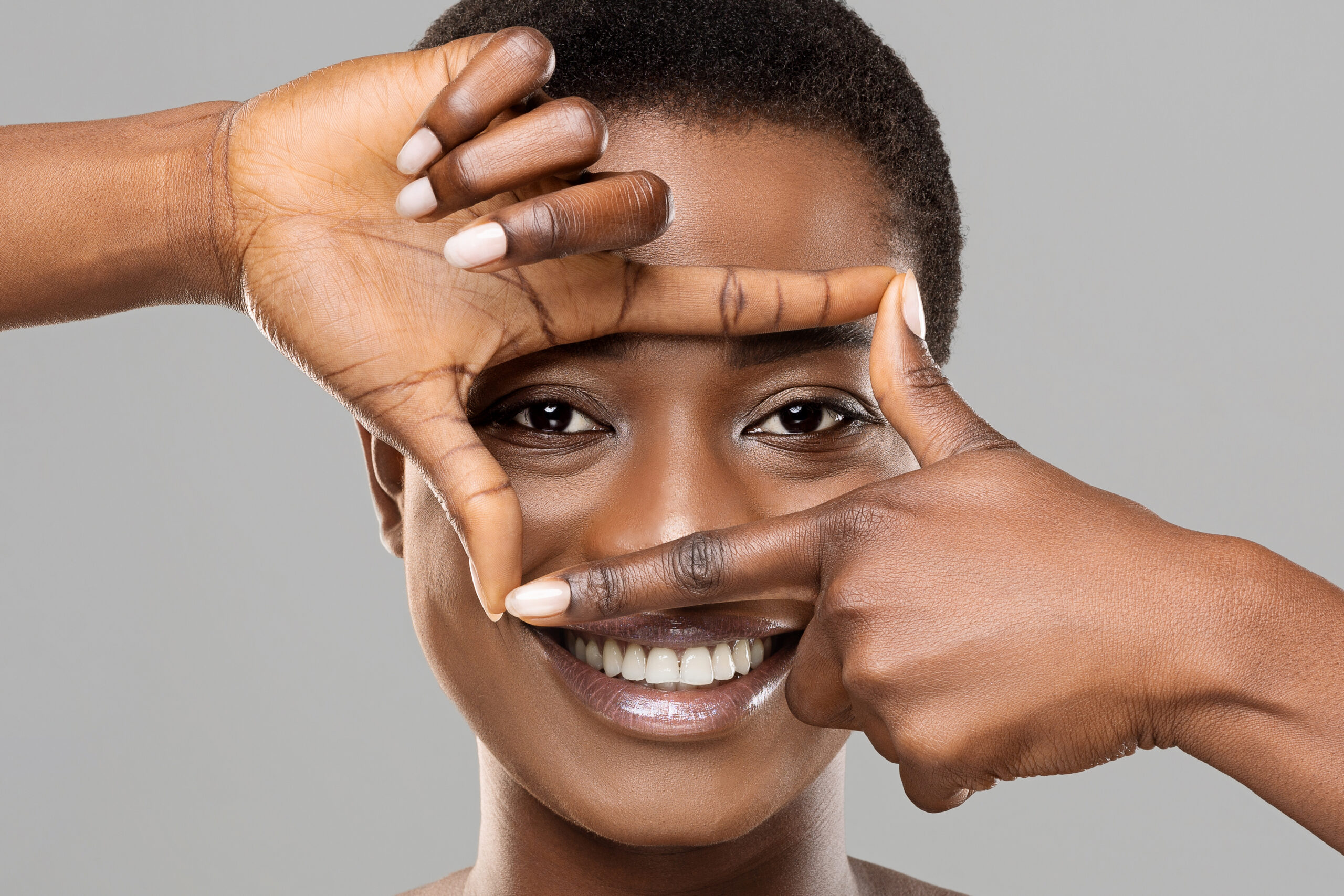 Smiling woman framing her face with her hands, showcasing natural beauty and healthy skin, close-up on face and hands, gray background.